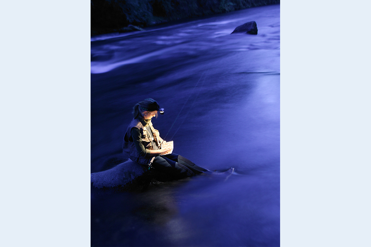 Waiting for sunrise a girl sits on a rock in a river picking out a fishing fly by headlamp light.
