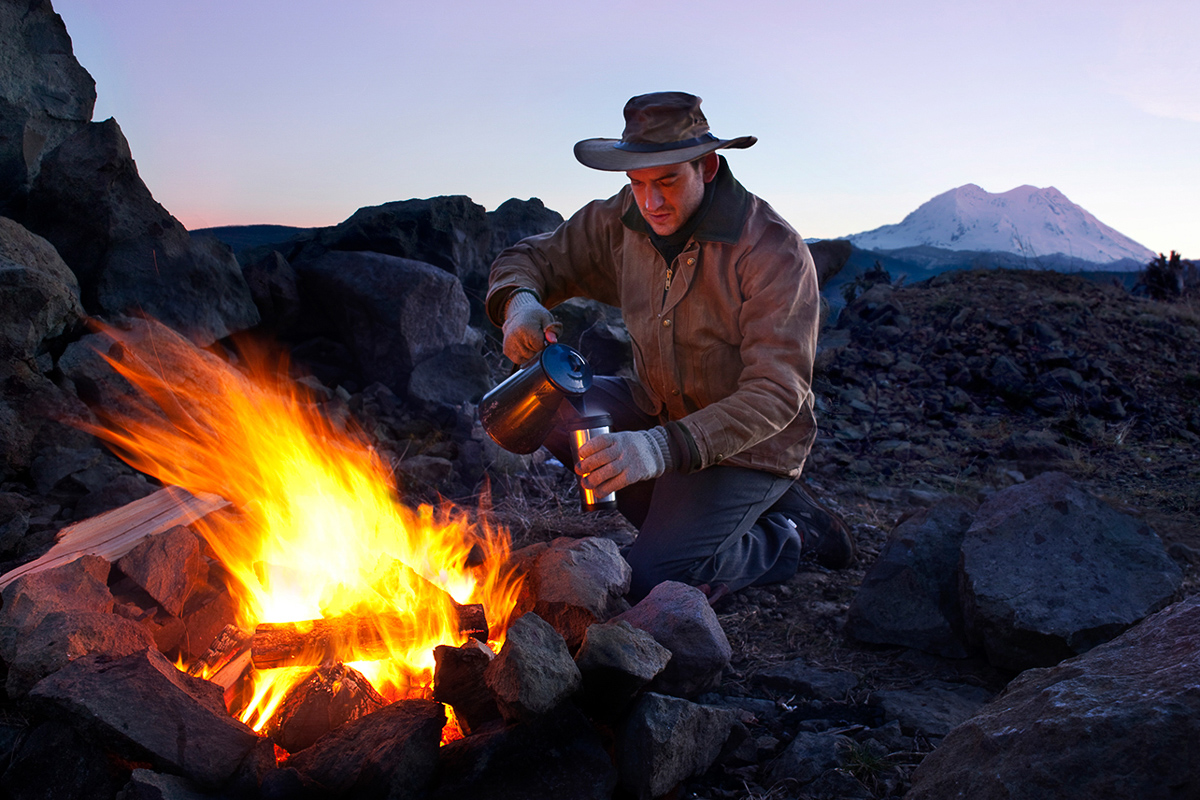 An outdoorsman pours a cup of hot coffee beside a blazing campfire with the sun rising over Mt.  Rainer in the background.